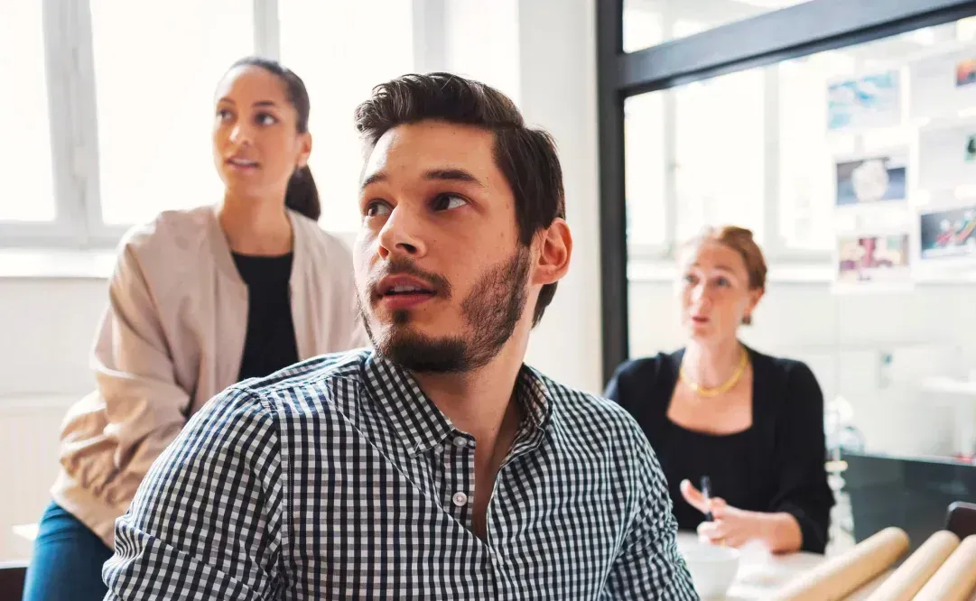 Man in plaid shirt looks to the side, with two women in the background in an office setting.
