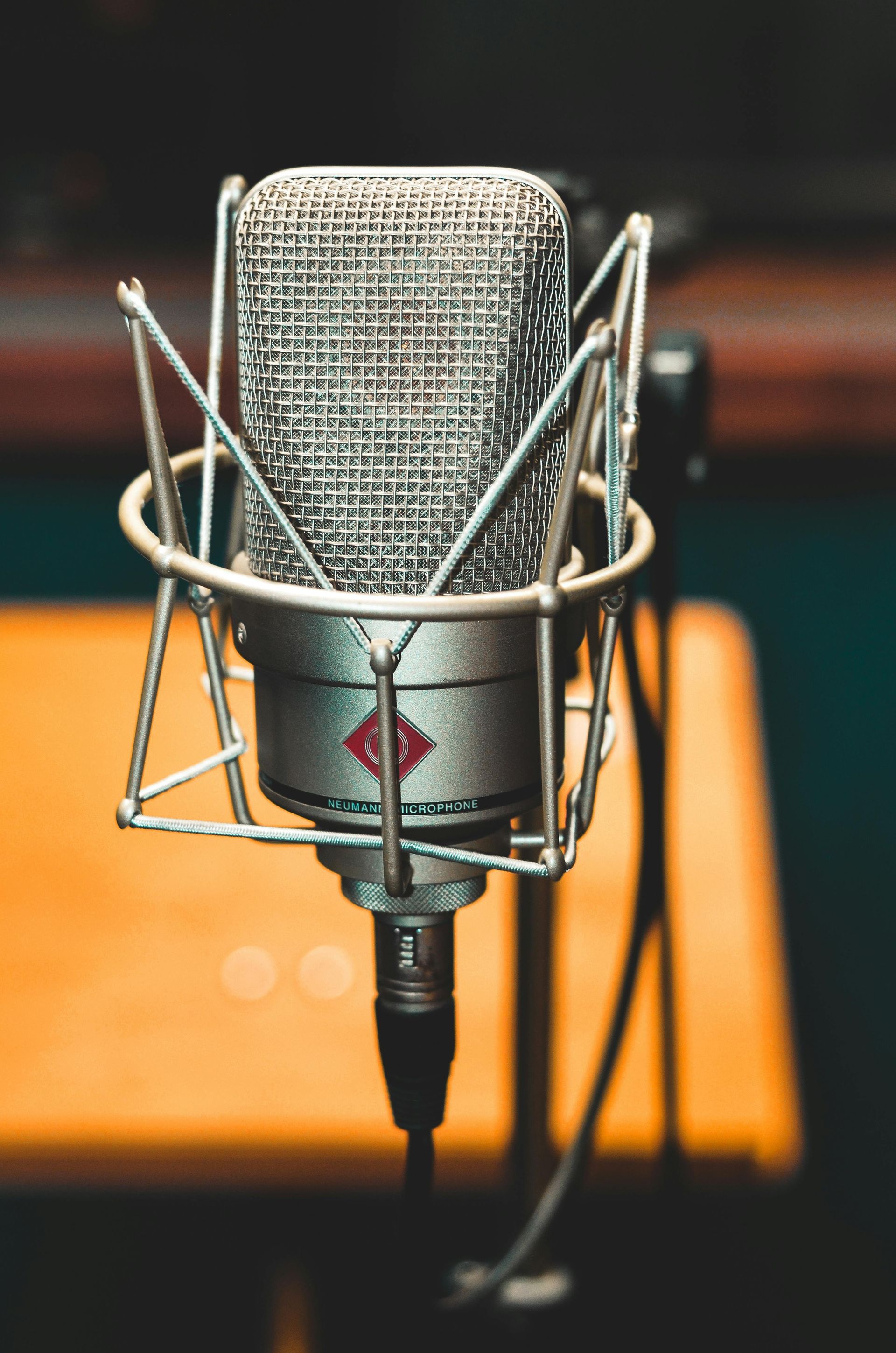 Silver microphone in a shock mount, close-up, with a blurred orange desk and background.