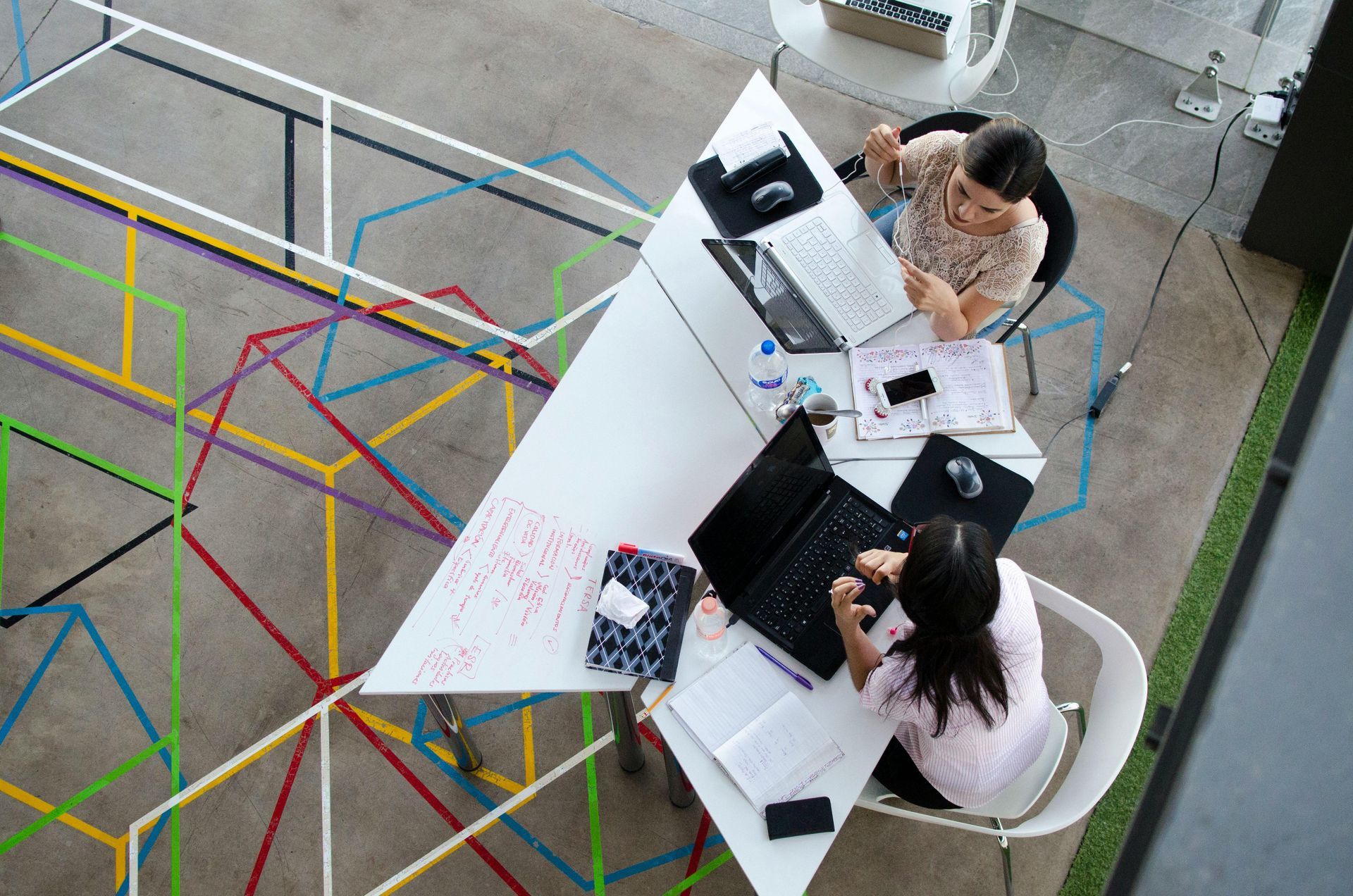 Two women working together at a white triangular table with laptops. Colorful geometric shapes on carpet.