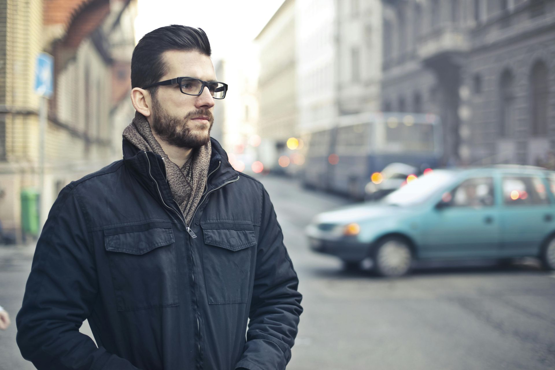 Man with glasses and beard on a city street, wearing a dark jacket and scarf, looking to the side.