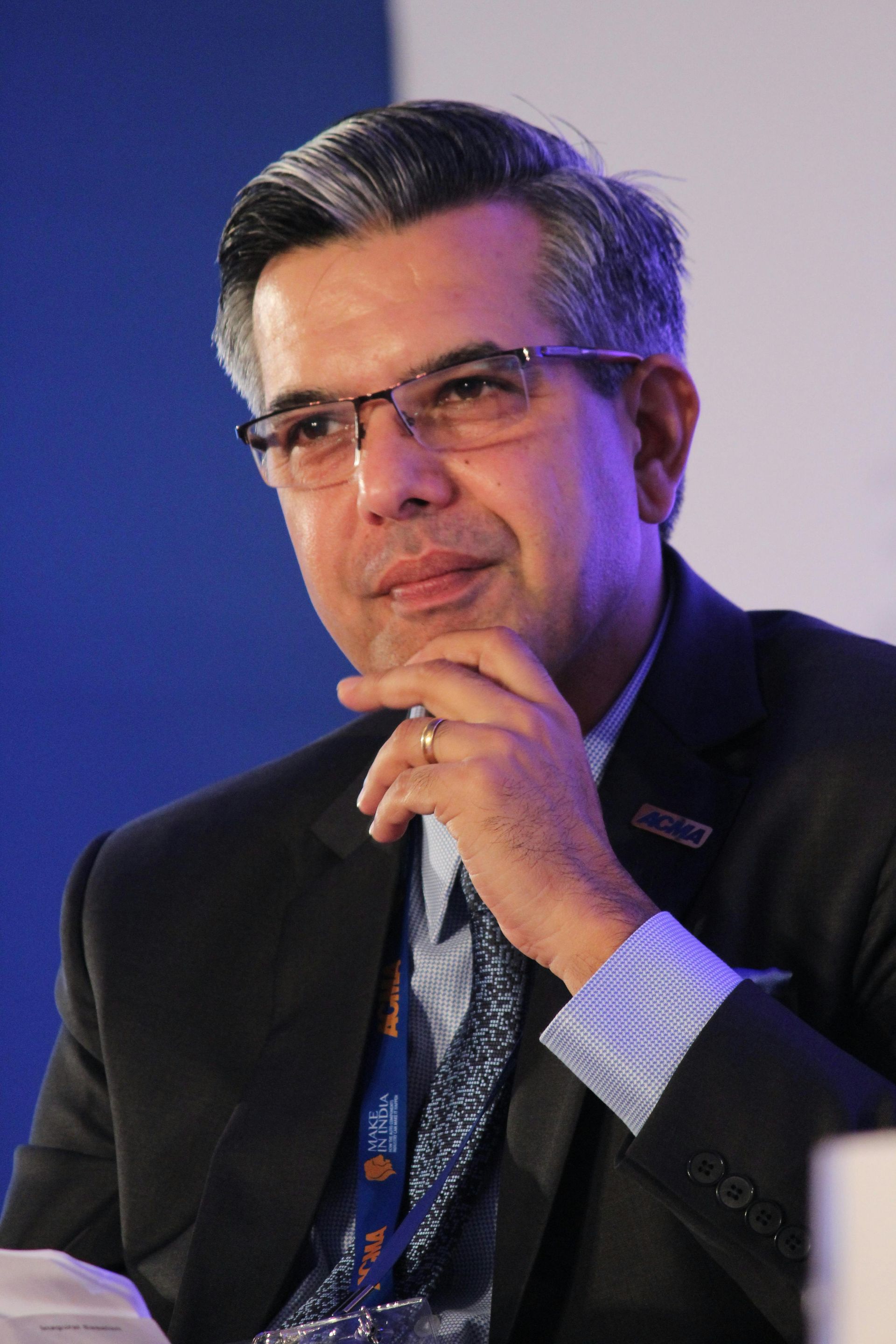 Man with glasses, hand to chin, wearing a suit and tie, speaking at an event.