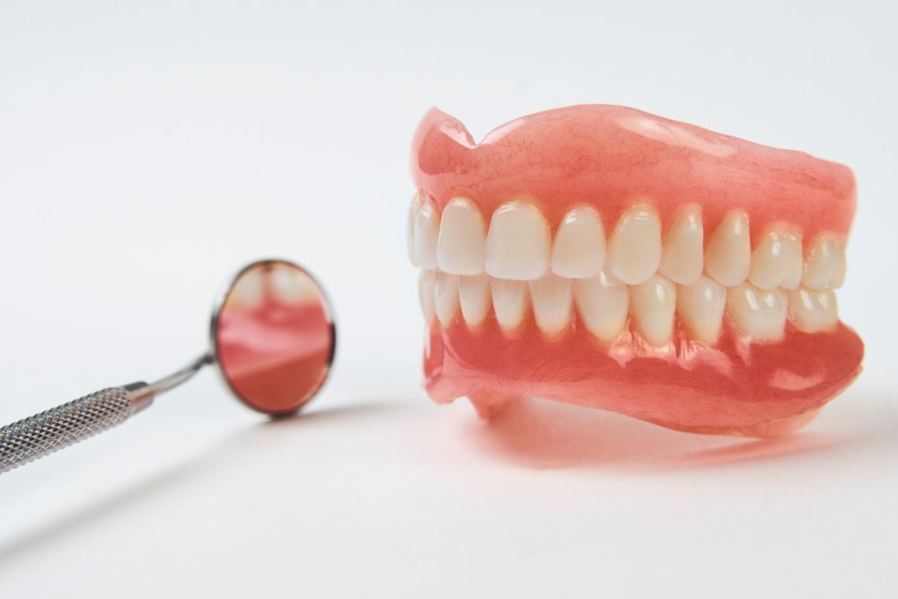A Denture And A Dental Mirror On A White Surface — Dentures Direct Taree-Rod Hinton In Port Macquarie, NSW