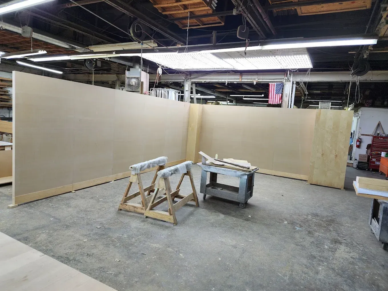 Workshop interior: large plywood walls in progress, saw horses, tools, and an American flag visible.