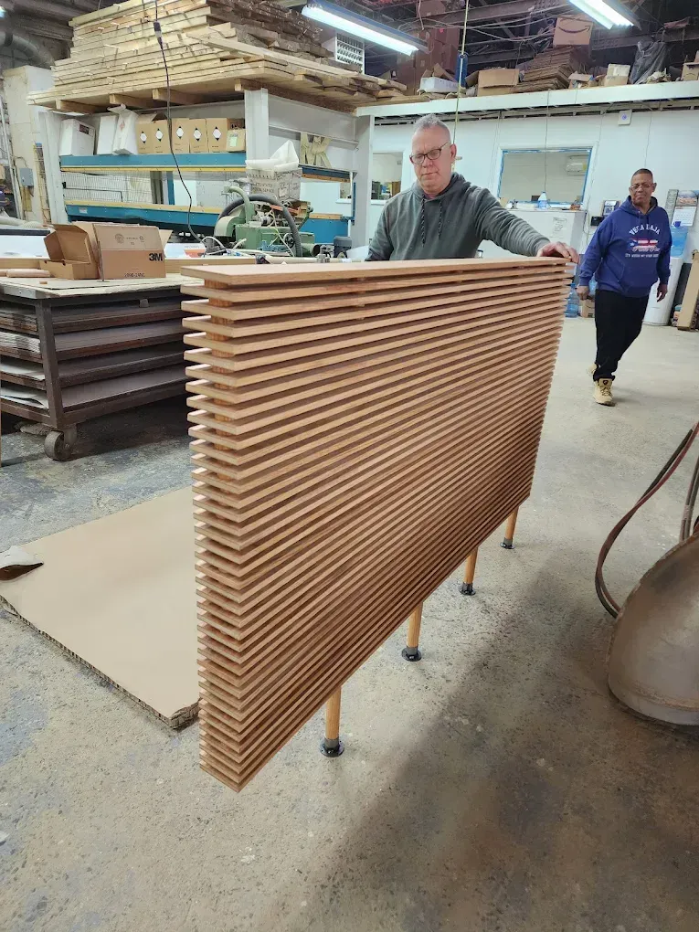 Man holding a large stack of wooden panels in a workshop. Another man walks in the background.