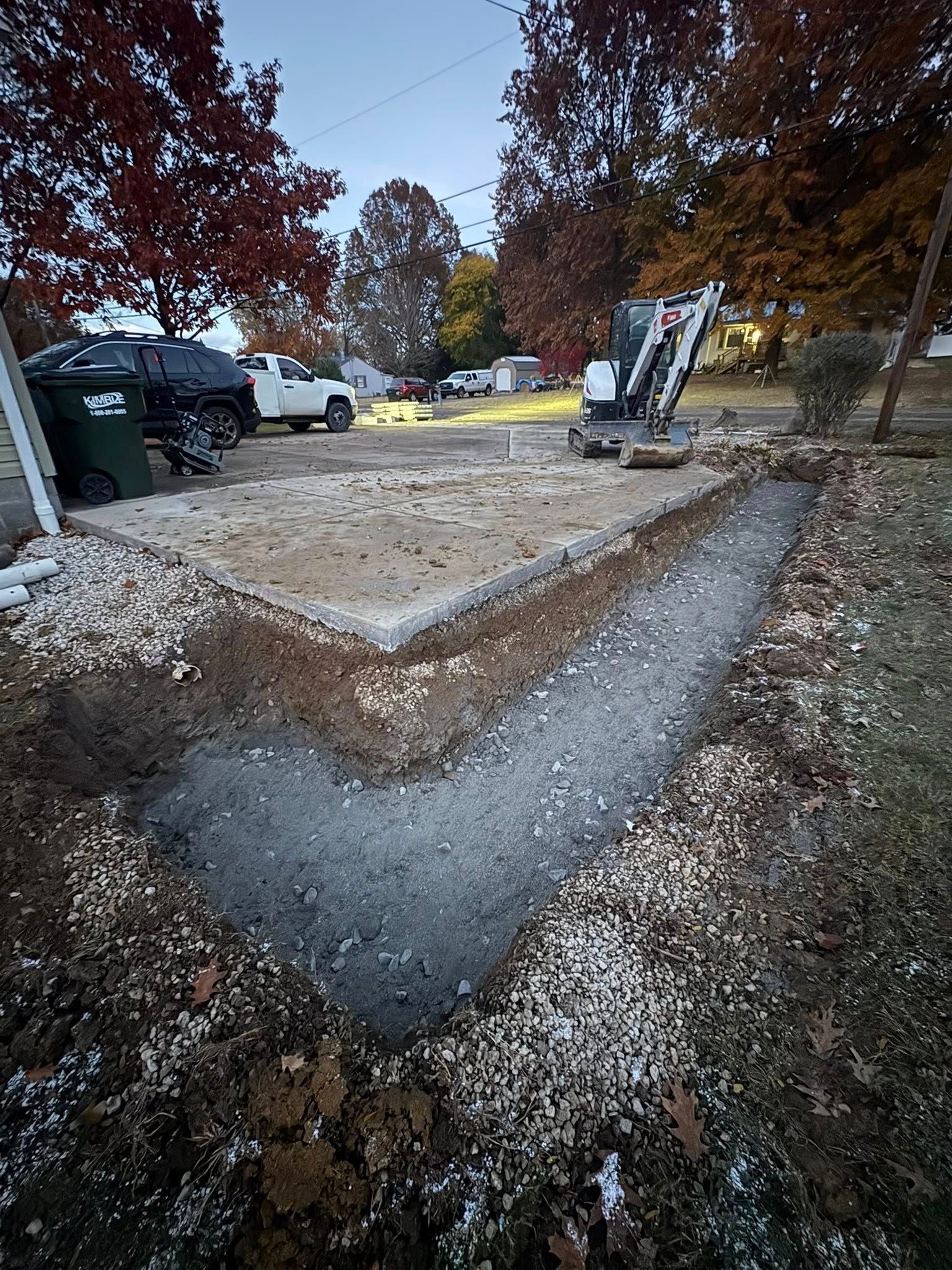 Freshly poured concrete walkway beside a driveway with trees, trash bins, and parked vehicles at dusk