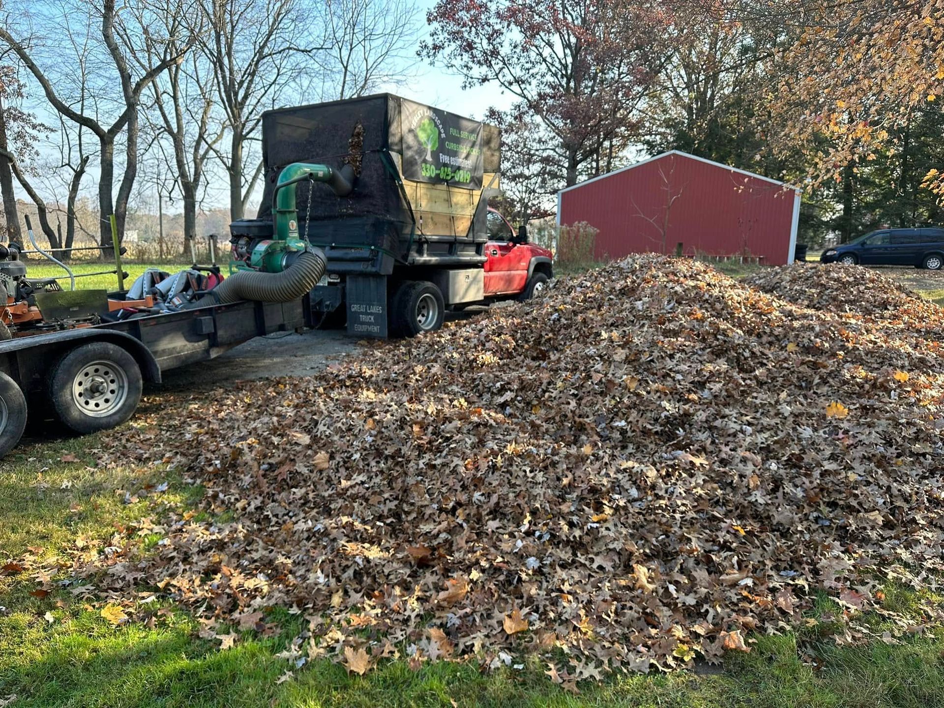 A truck unloading a large pile of leaves in a wooded yard beside a red shed.