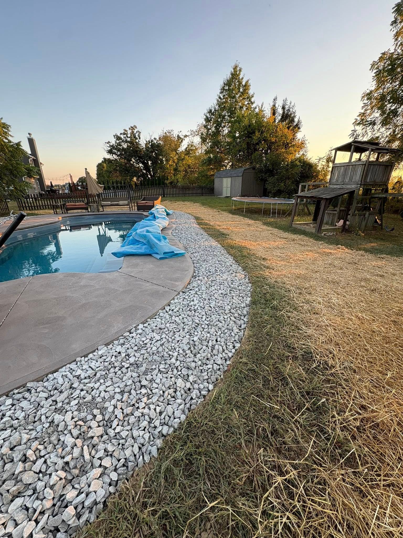 Backyard pool at sunset with white gravel border, lawn, trees, and a covered blue object near the edge