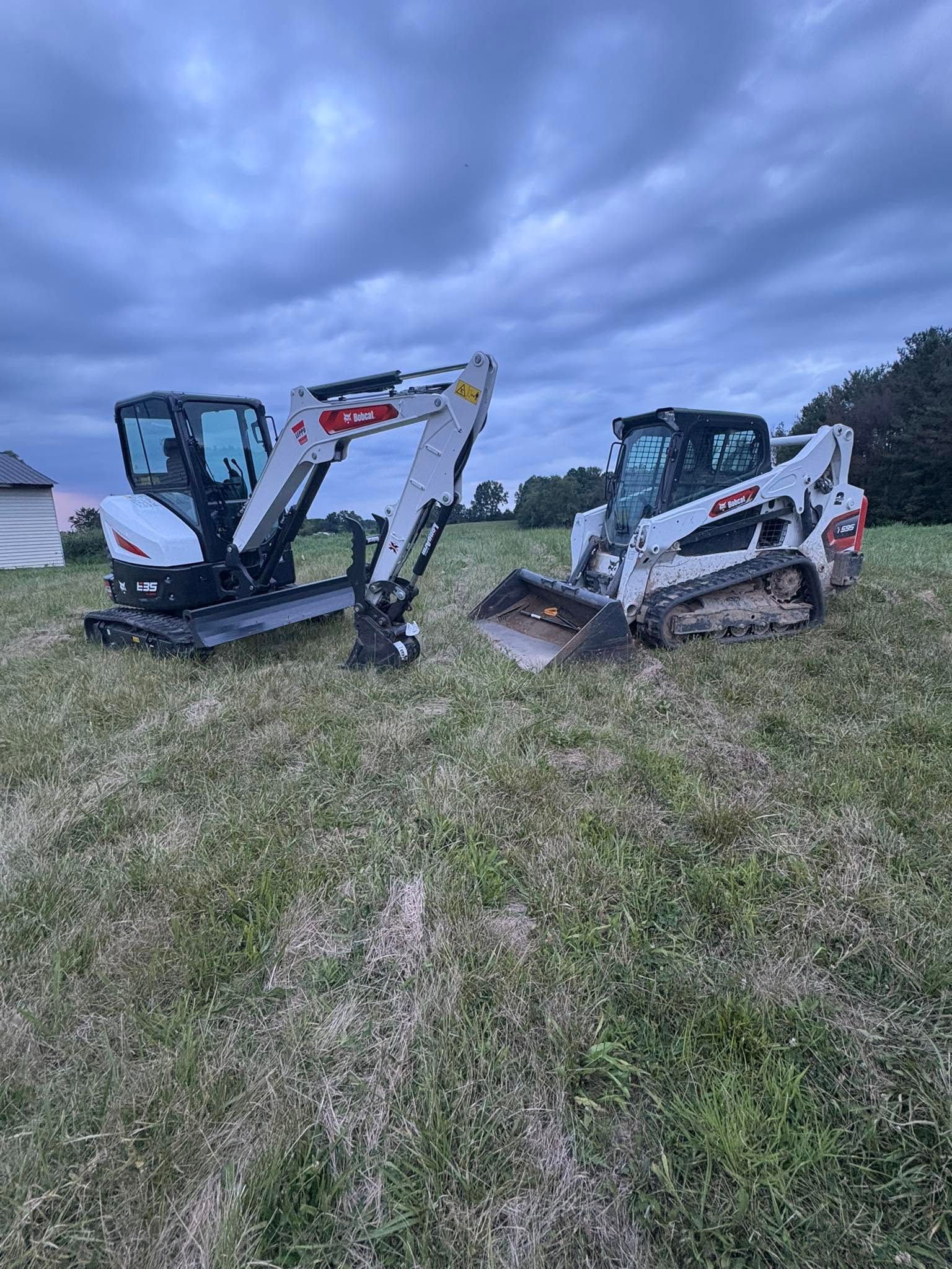 Two mini excavators on a grassy field under a cloudy sky, with a small bucket attachment between them