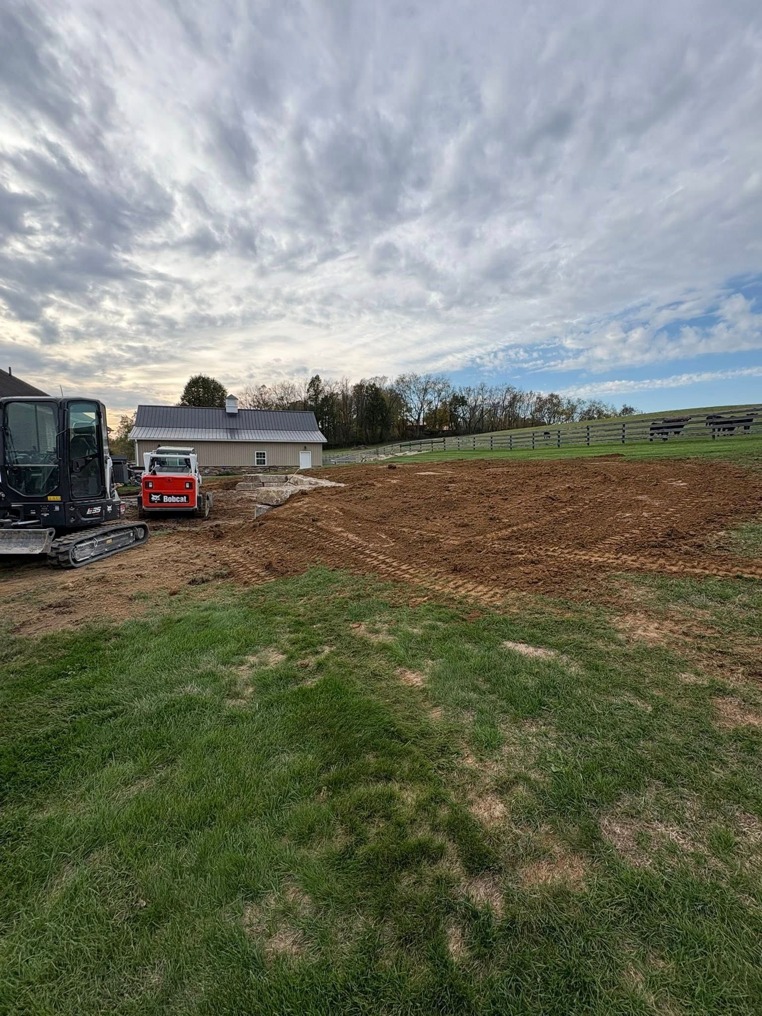 Construction site with excavator, red truck, and dirt lot beside a house under cloudy sky