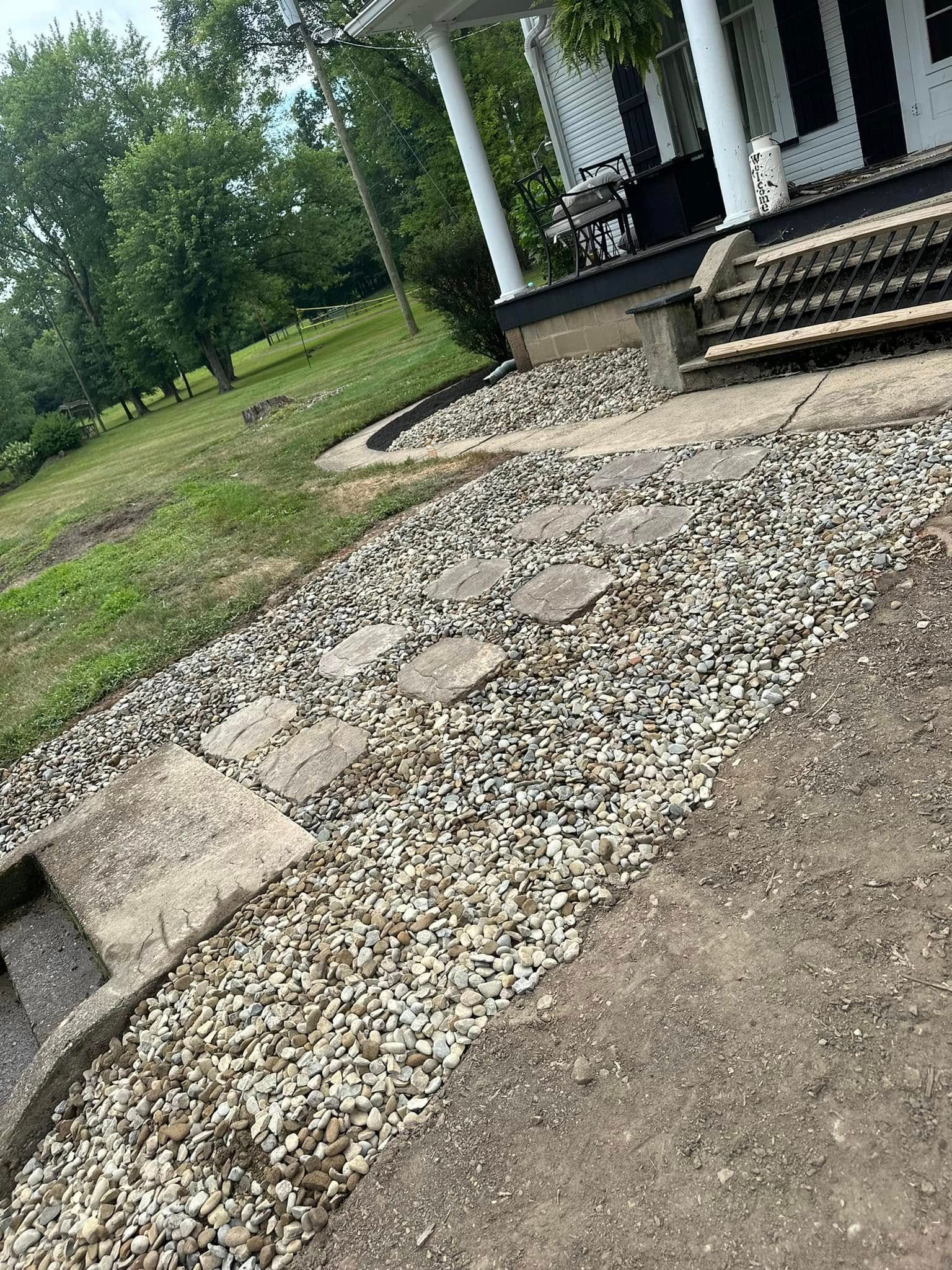 Gravel walkway beside a house porch with steps, concrete edges, and a grassy yard nearby
