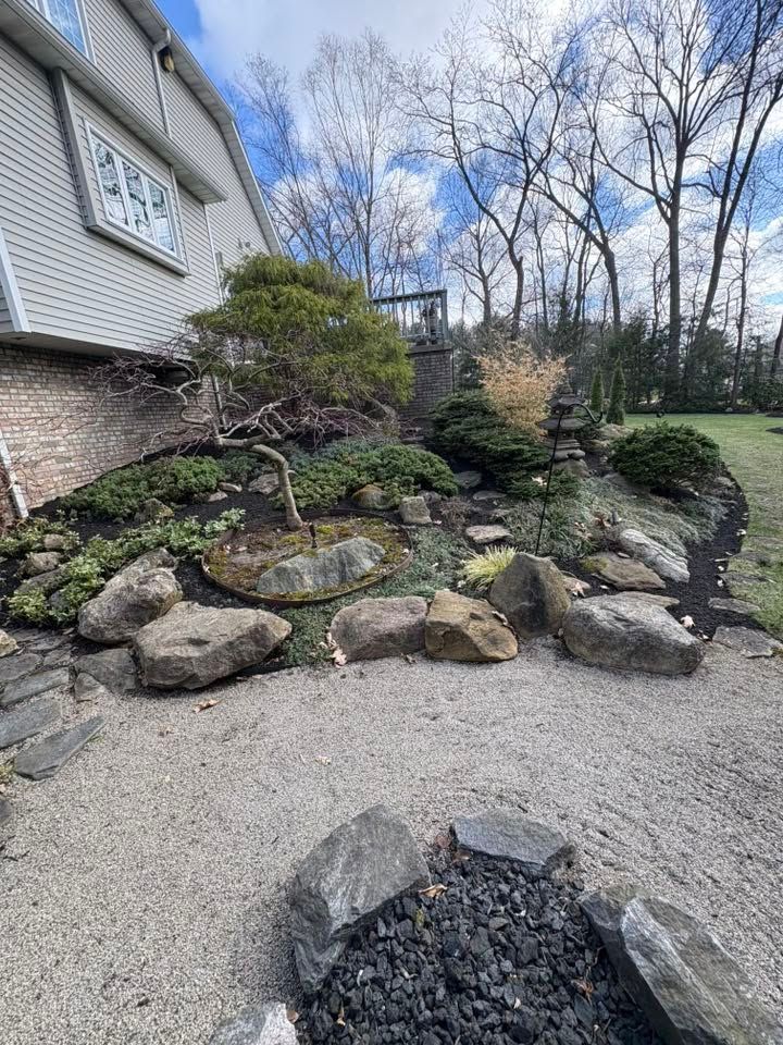House side yard with rock garden, evergreen shrubs, and gravel driveway under bare trees