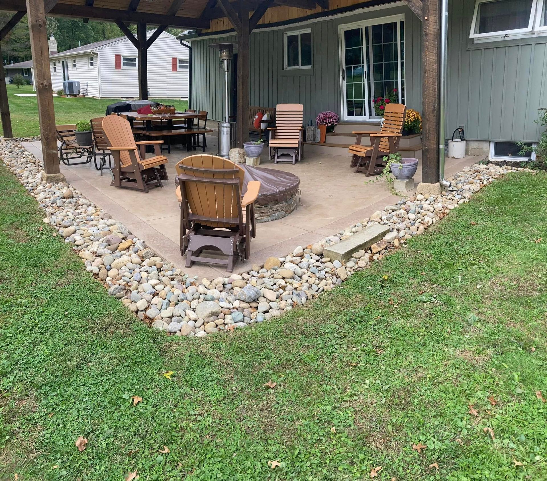 Covered backyard patio with wicker chairs and a fire pit, bordered by decorative rocks and grass.