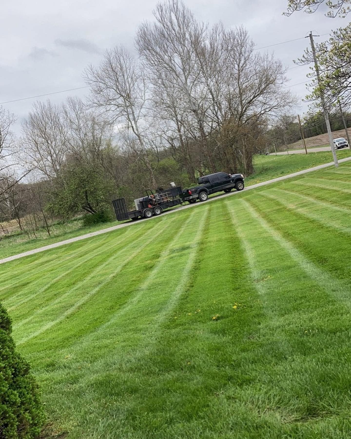 Black pickup and trailer on a mowed grassy field near leafless trees under a cloudy sky
