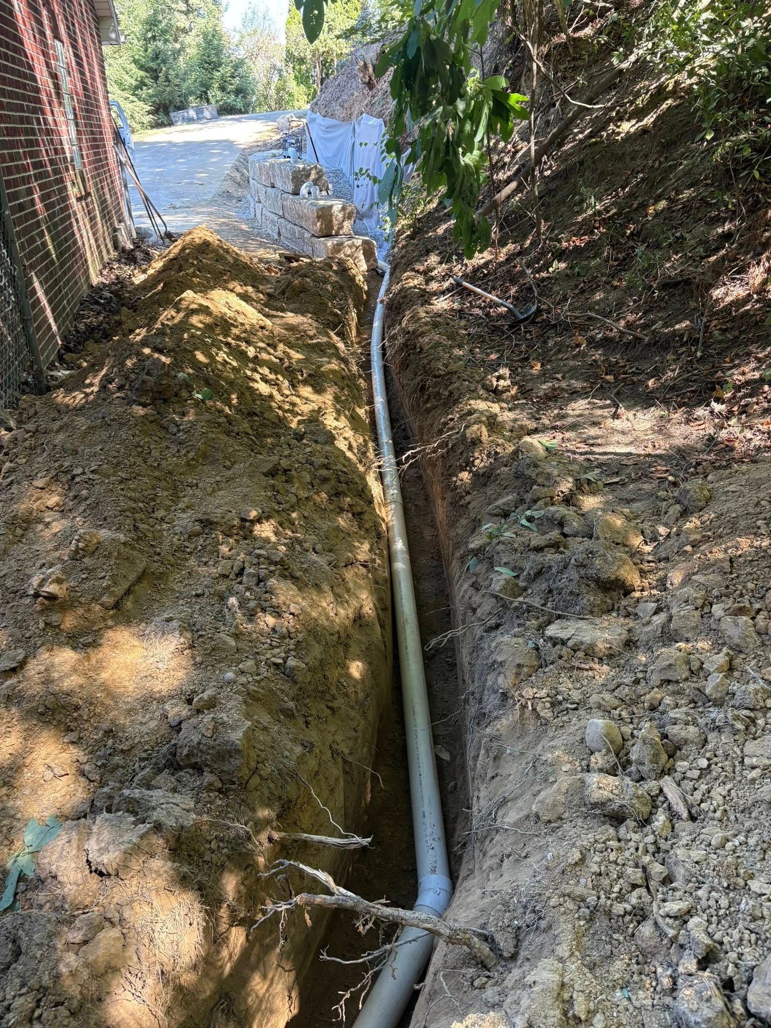 Narrow trench with a white pipe running through dirt between fences and trees near water.