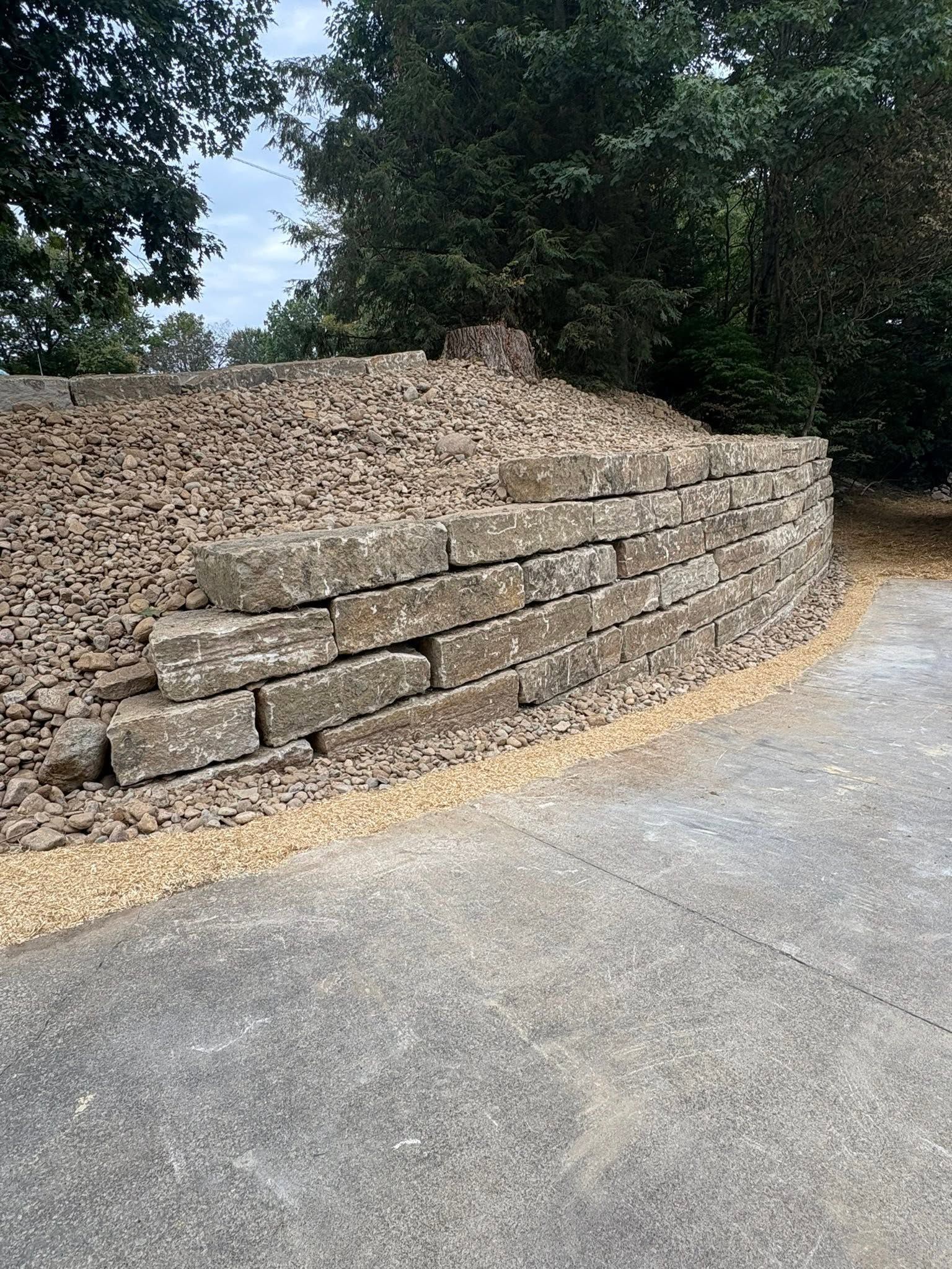 Stone retaining wall beside a road, topped with mulch and bordered by trees