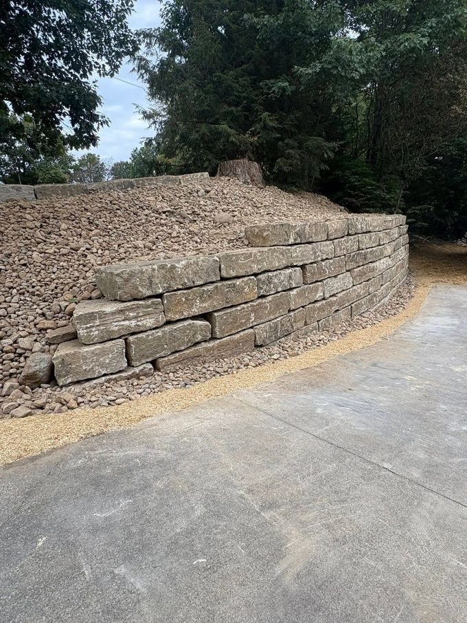 Retaining wall of stacked stone blocks beside a paved road, topped with tan gravel and surrounded by trees.