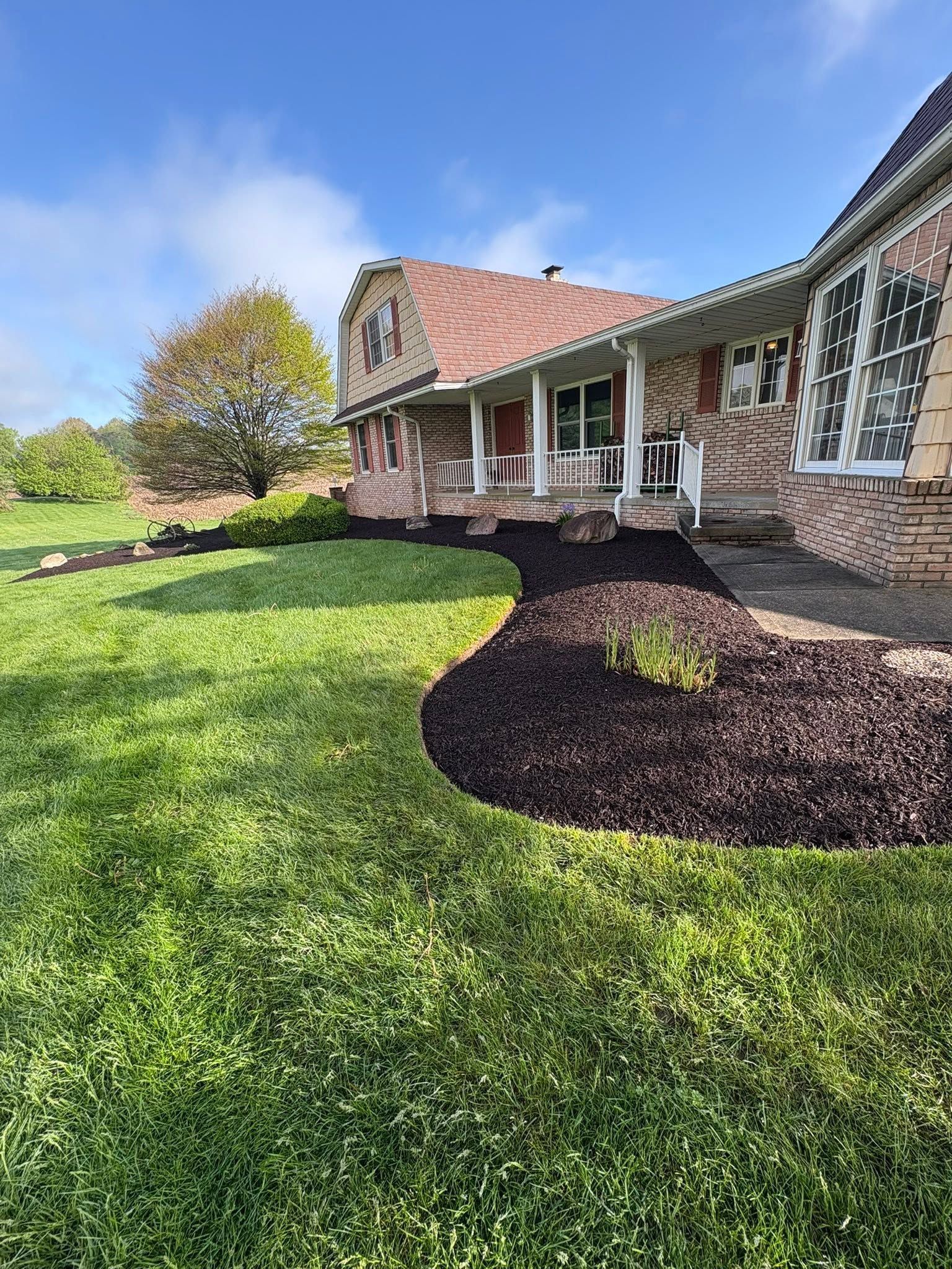 Brick house with porch and landscaped yard, featuring a curved mulch bed and green lawn under a blue sky