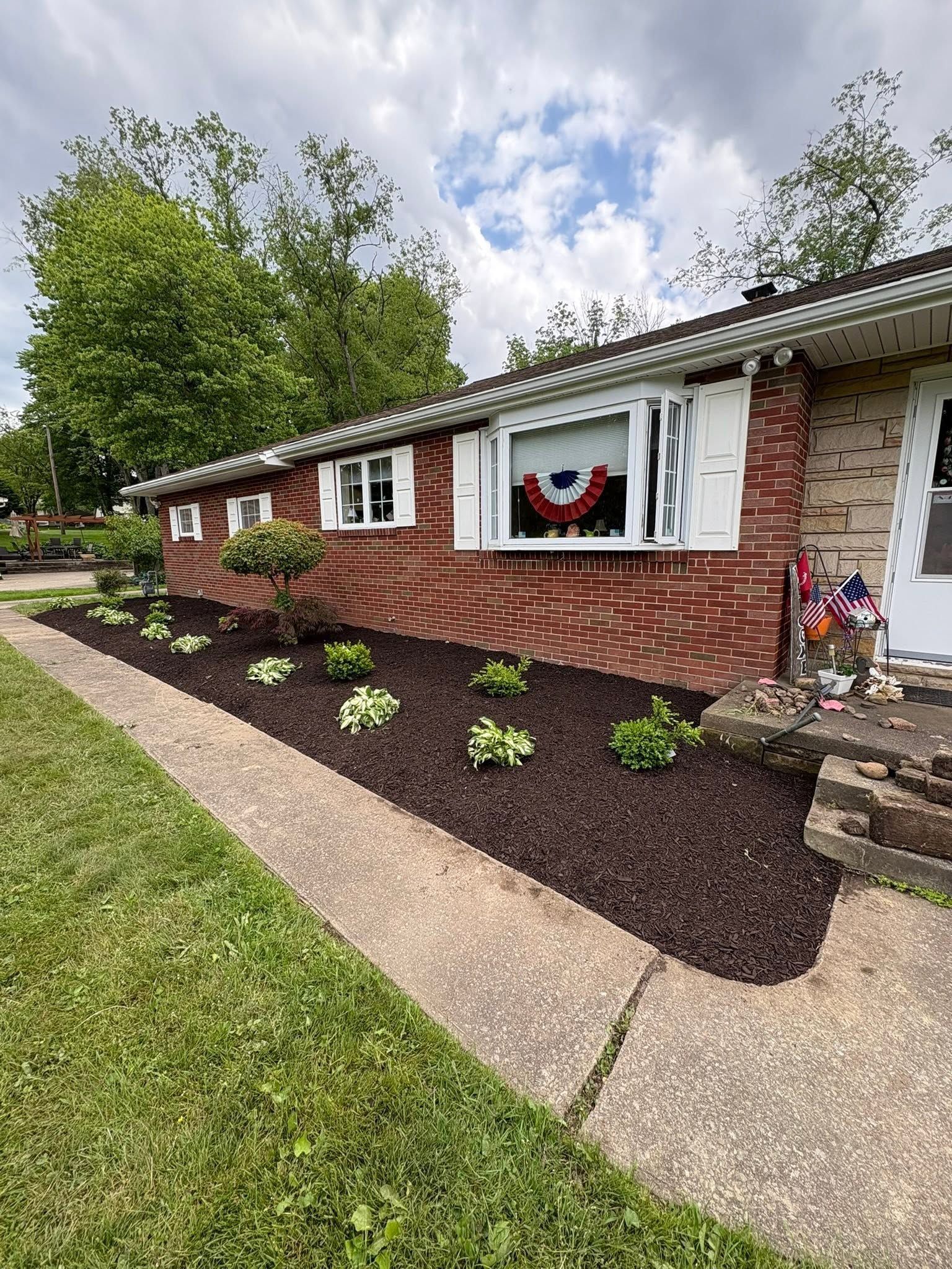 Brick ranch house with patriotic window decor, fresh mulch beds, and small shrubs along a concrete walkway