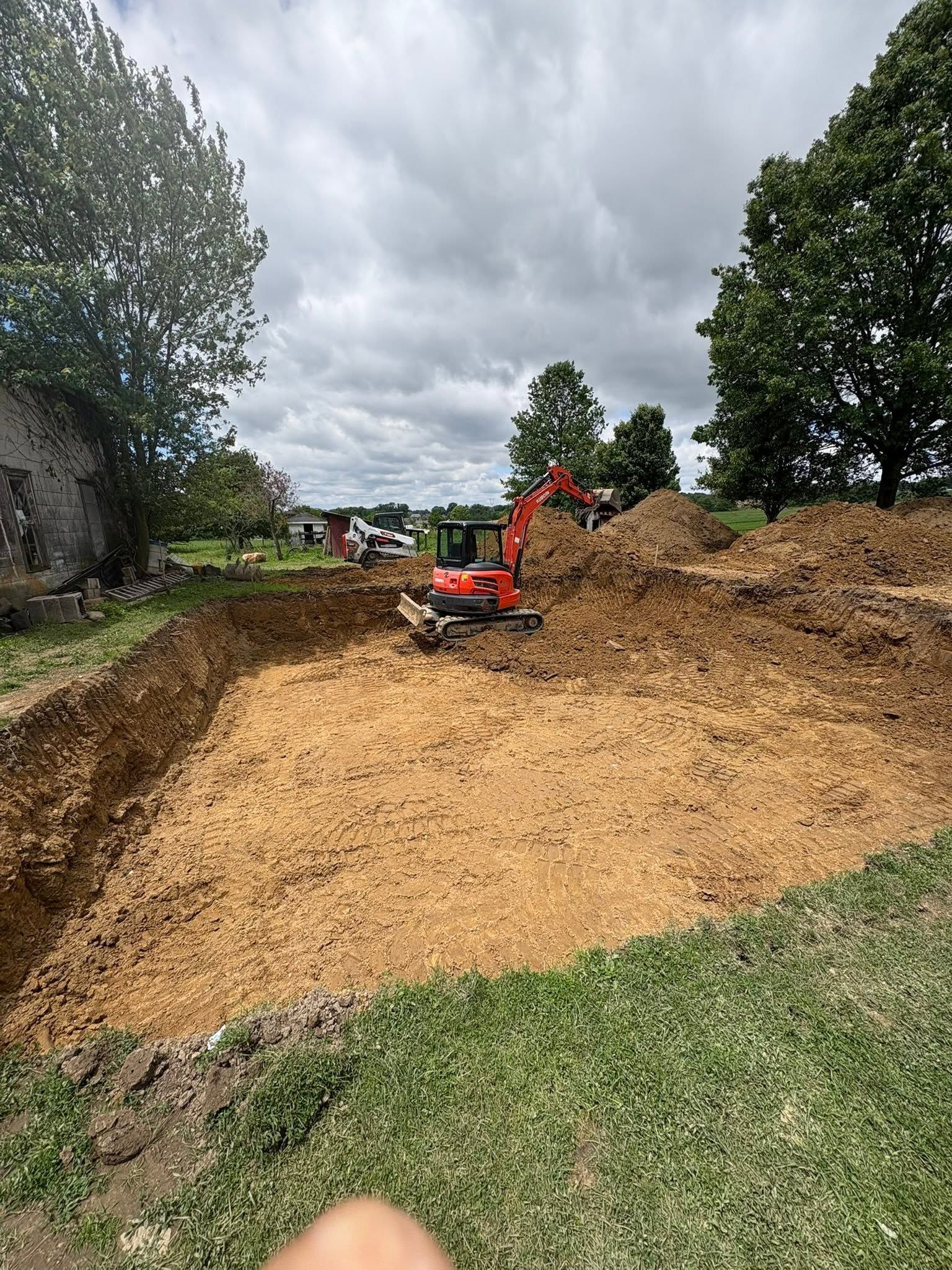 Orange excavator clearing a dirt embankment on a cloudy construction site with trees nearby