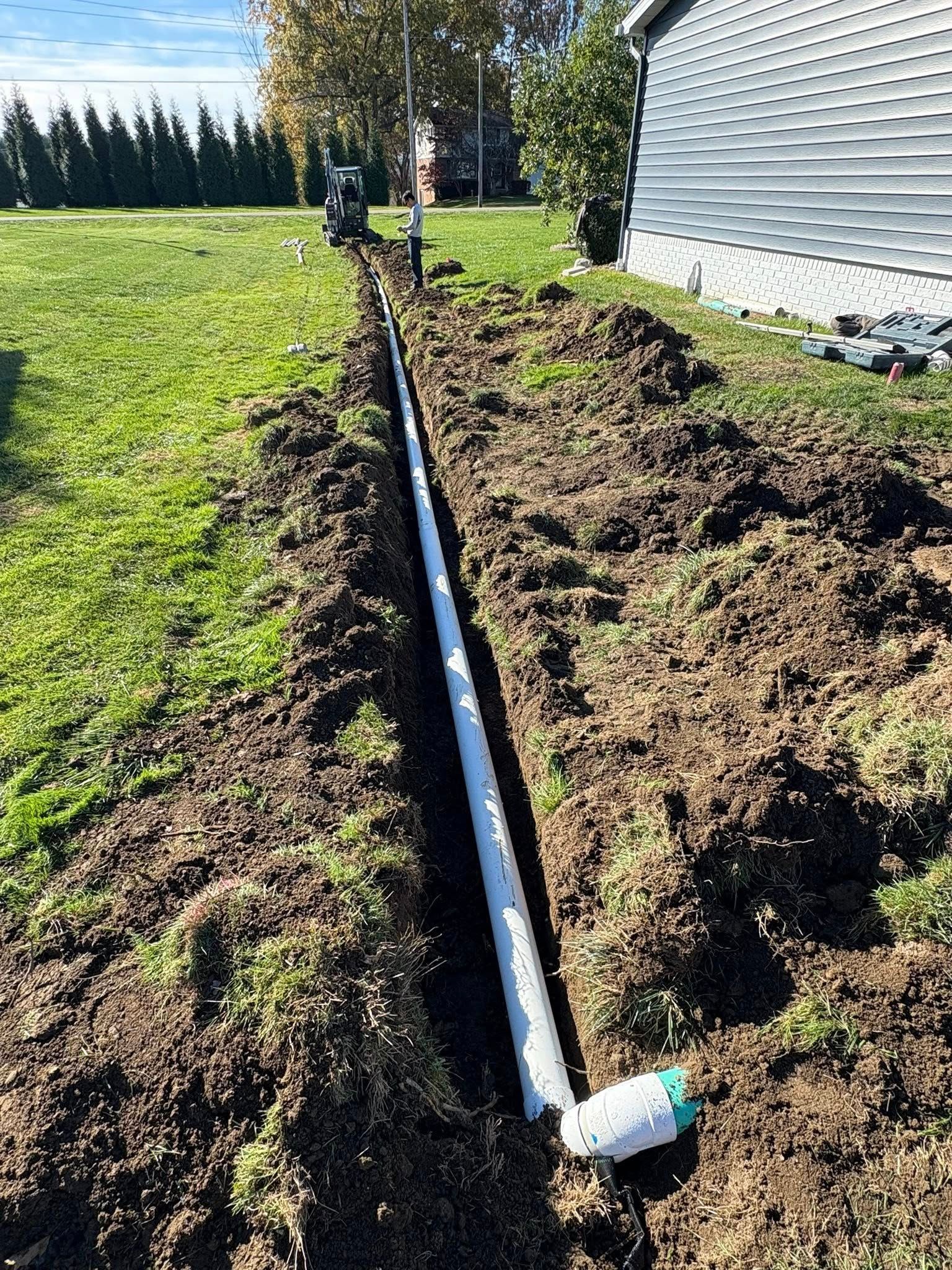 Trench beside a house with a long black pipe laid in it and fresh dirt piled along the edge
