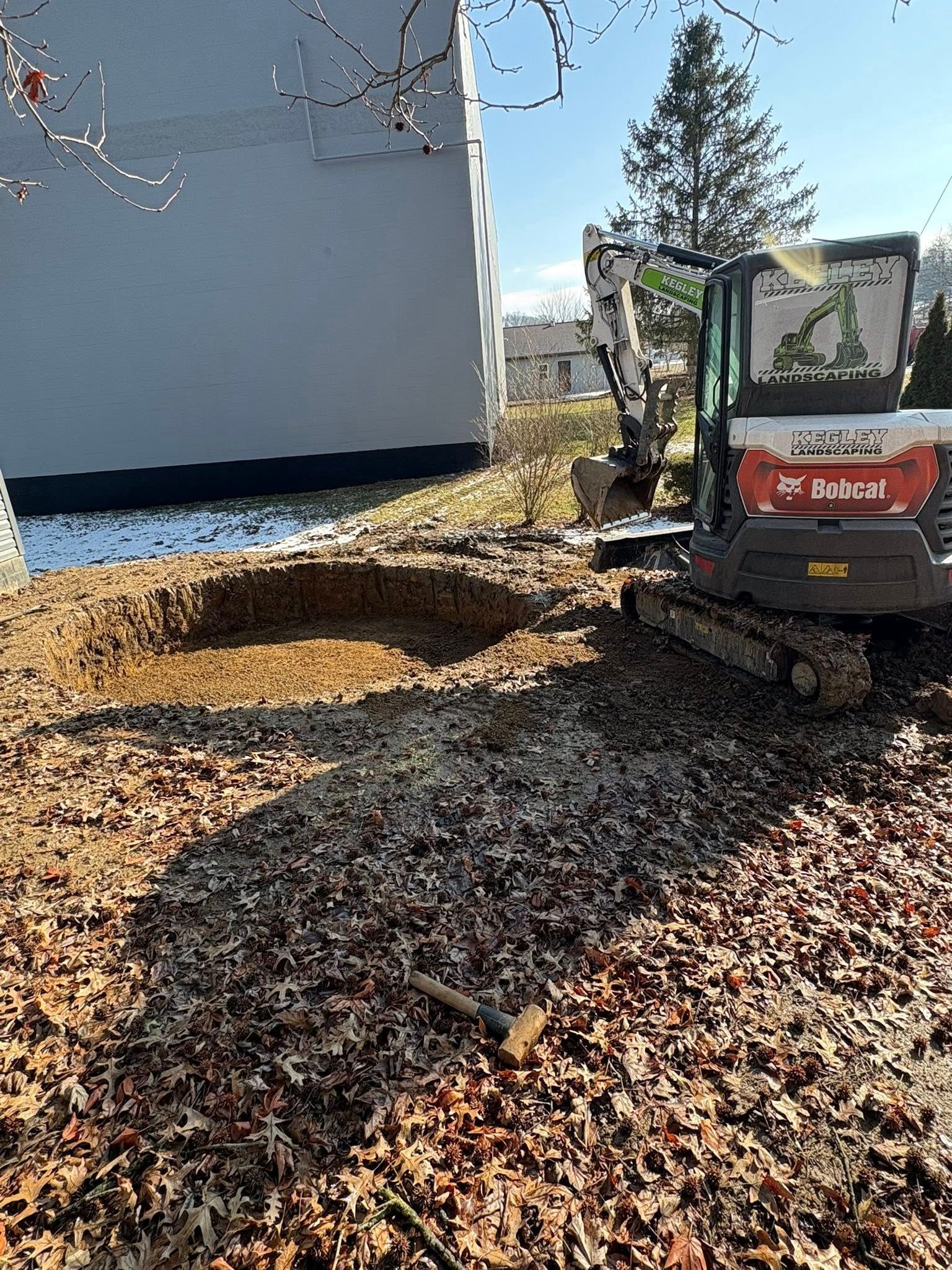 Small excavator beside a house, digging a dirt mound under bare trees on a sunny day