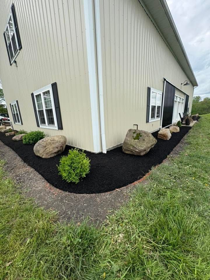 White metal-sided building corner with black mulch beds, rocks, shrubs, and windows along a grassy edge