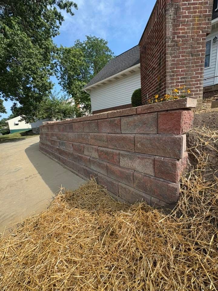 Low brick retaining wall beside a driveway, with straw mulch and a brick house corner in daylight