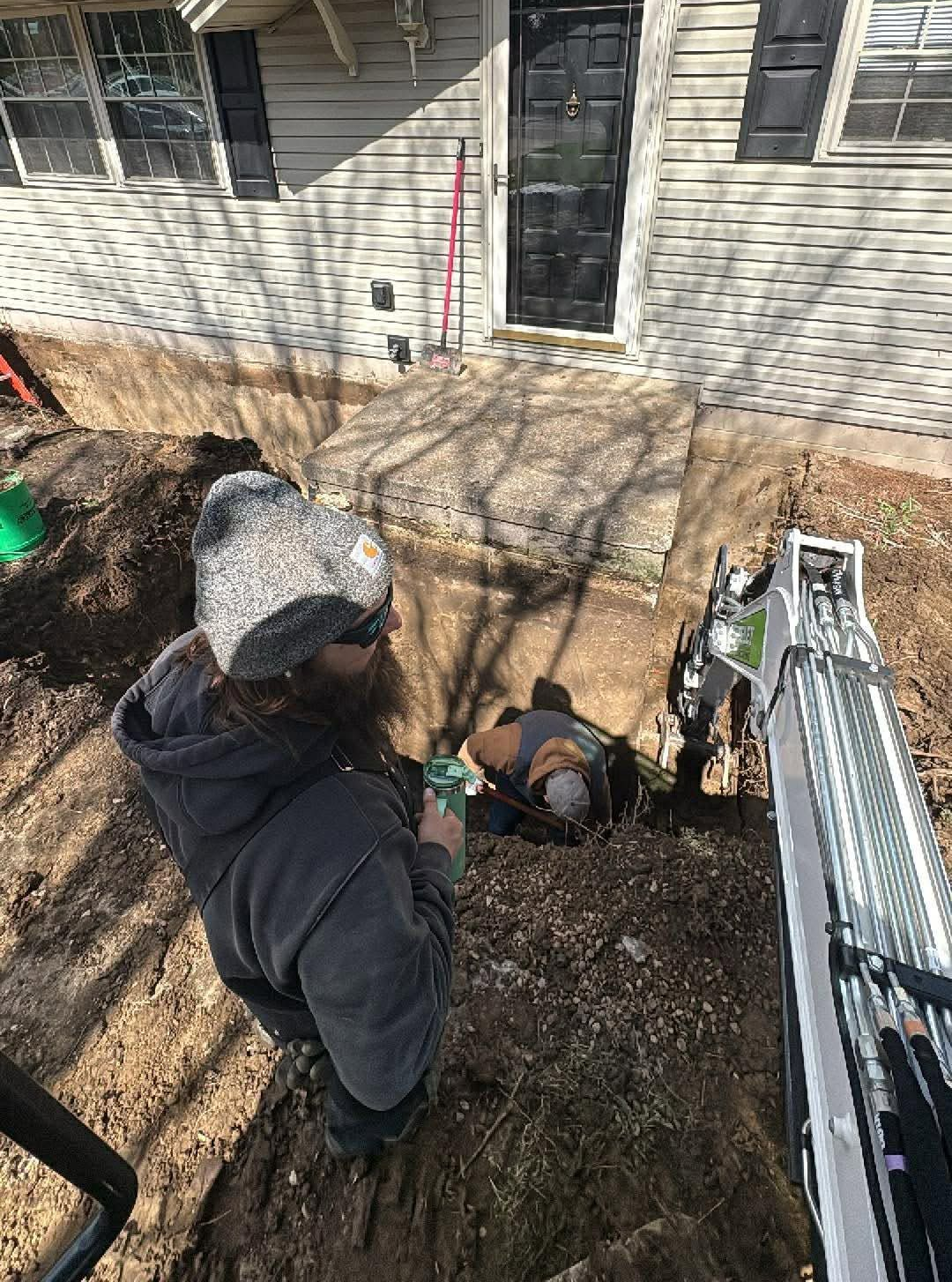 Two workers digging a trench beside a house, with one standing and one in the hole.