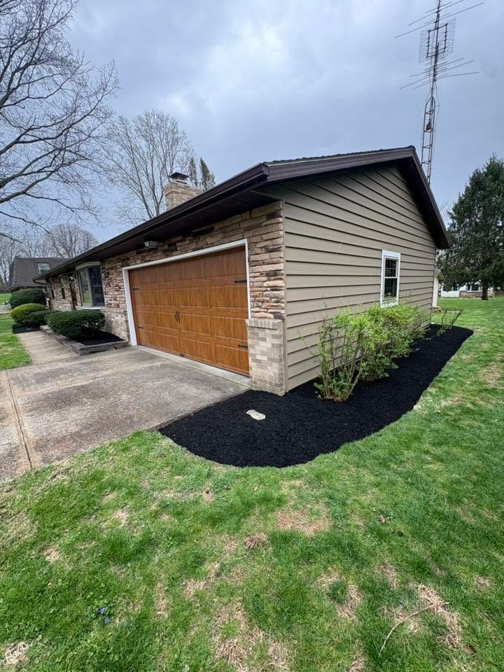 Suburban garage with wood doors, beige siding, and black mulch landscaping on a cloudy day