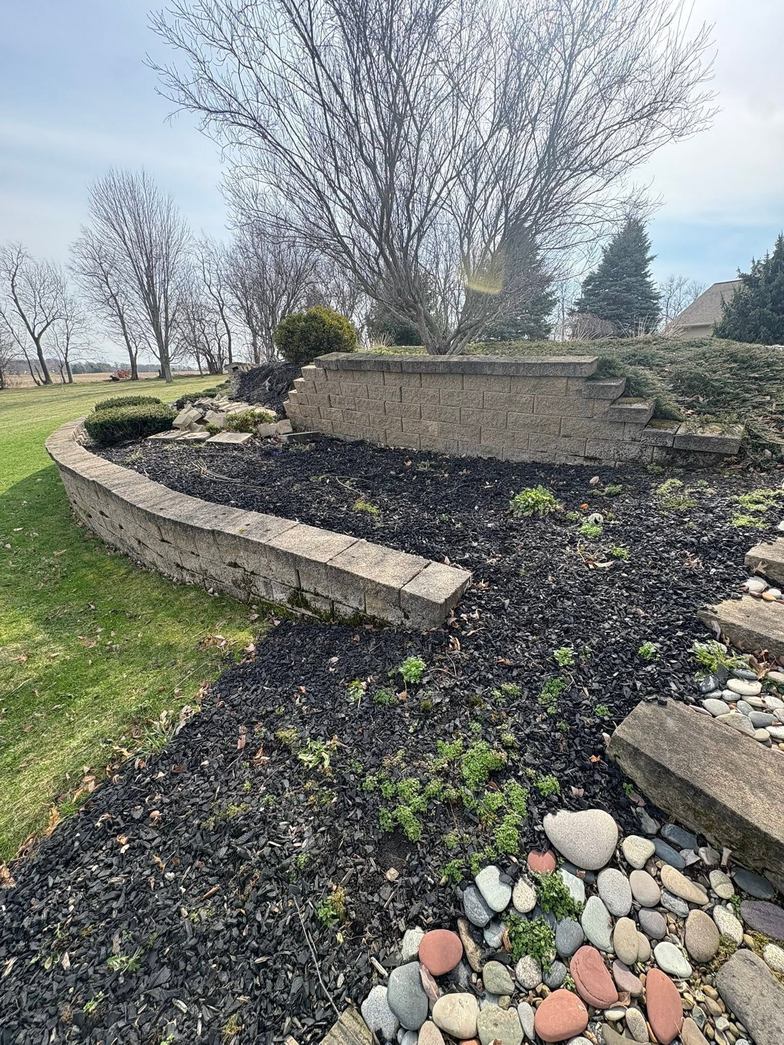 Curved stone garden wall with dark mulch, scattered rocks, and leafless trees on a grassy landscape.