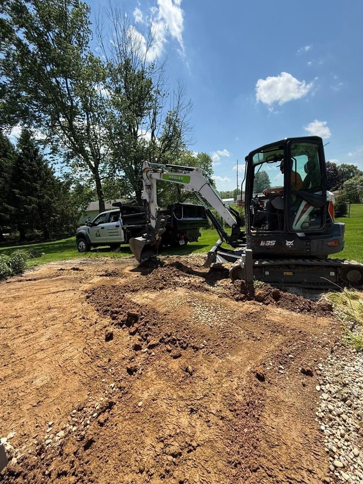 Compact excavator digging mulch in a sunny wooded yard beside a parked truck