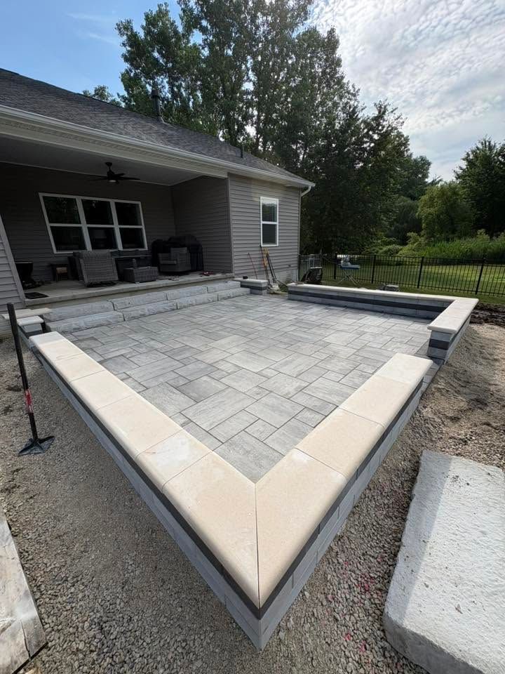 Raised stone patio under construction beside a house, with beige coping and a gray paver surface.