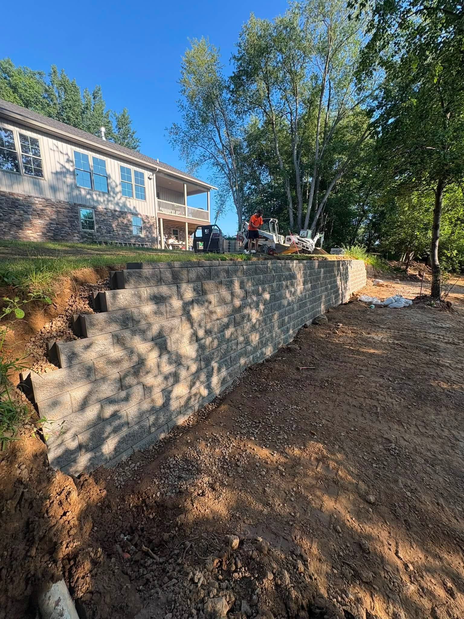 Stone retaining wall on a wooded slope beside a house, with trees and a person in the background.