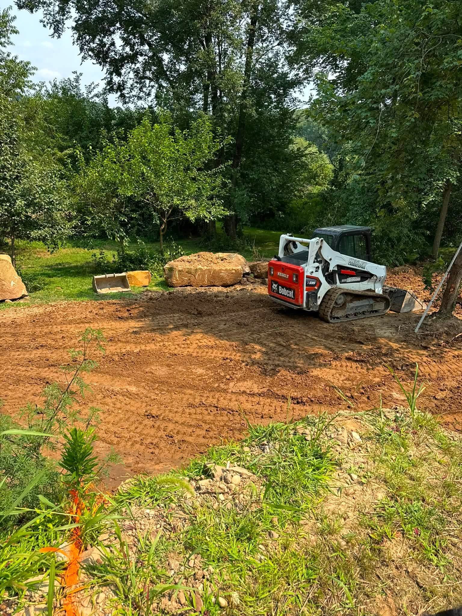 Small excavator leveling a dirt lot beside trees, with hay bales in the background.