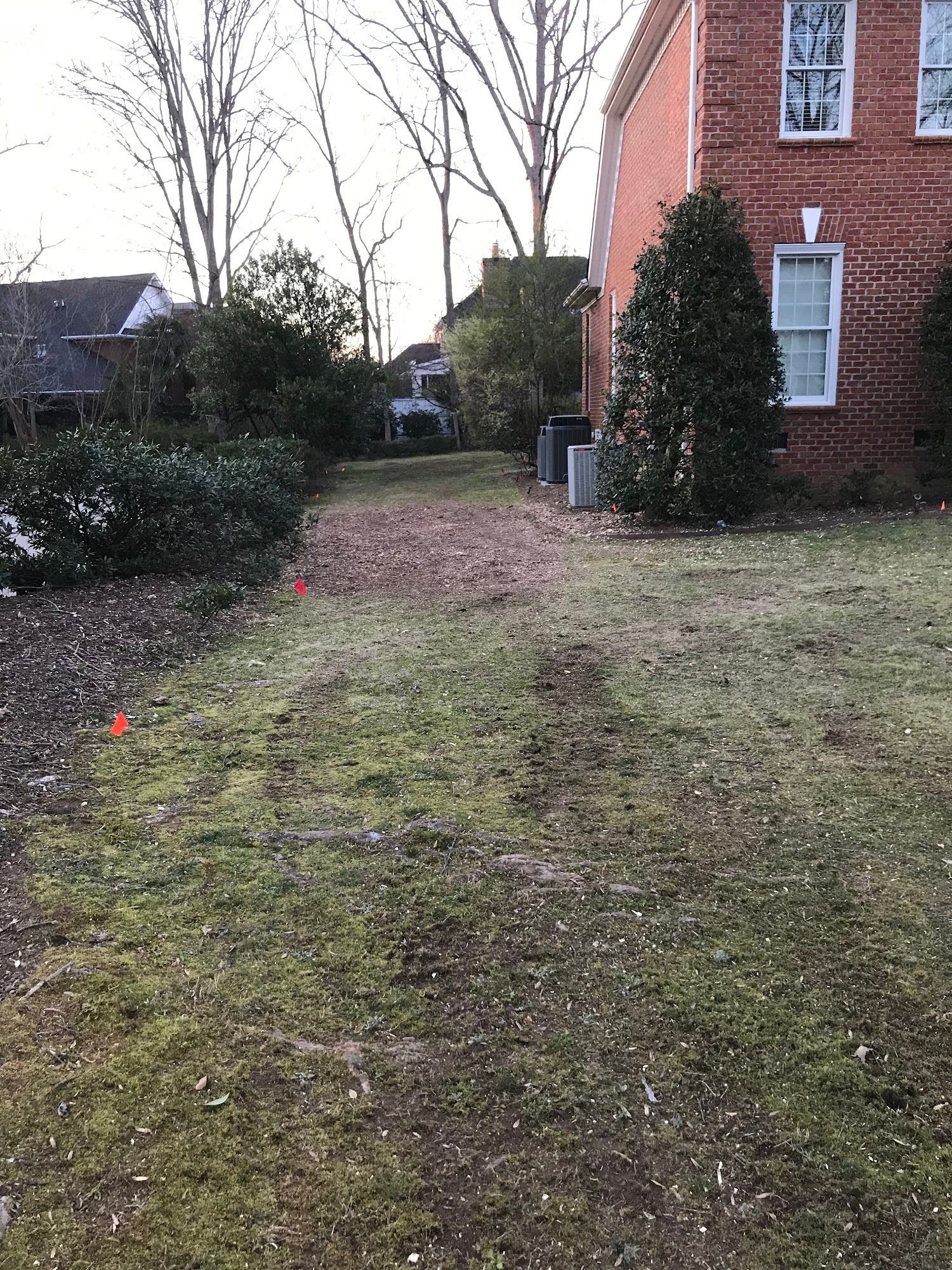 Narrow grassy yard beside a brick house, with shrubs, bare trees, and a path leading toward the back.