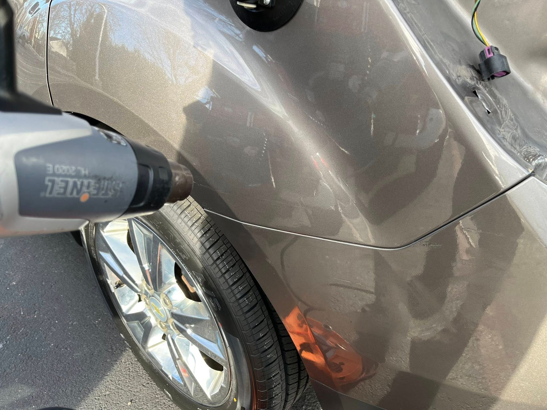 A hand uses a heat gun on the rear fender of a brown car to repair a dent.