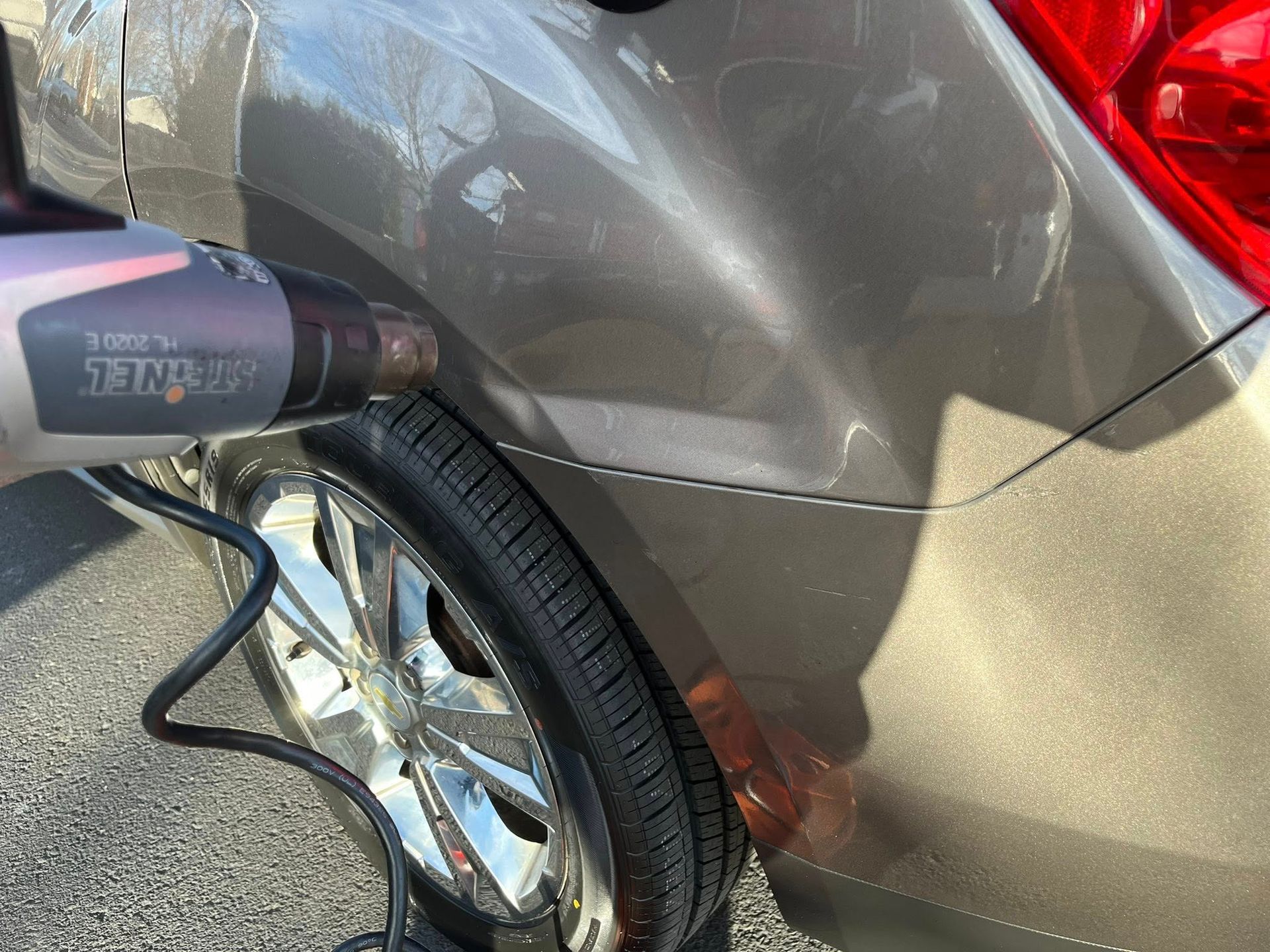 A heat gun being used to warm the dented rear fender of a brown car parked on asphalt.