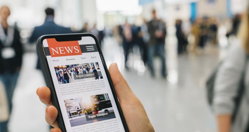 Eine Hand, die ein Smartphone hält, auf dem eine Nachrichtenwebsite zu sehen ist, in einem belebten öffentlichen Innenraum.
