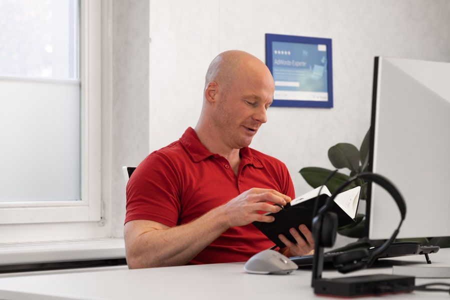 Mann im roten Hemd am Schreibtisch, schaut in ein Notizblock, Computer und Headset stehen vor ihm.