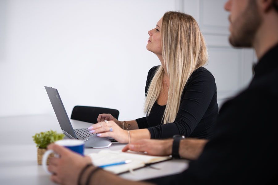 Frau sitzt an einem Tisch und benutzt einen Laptop, neben einer weiteren Person.