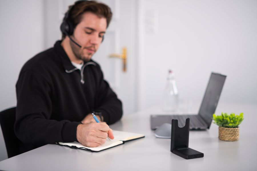 Mann mit Headset schreibt in Notizbuch, arbeitet am Laptop am Schreibtisch.