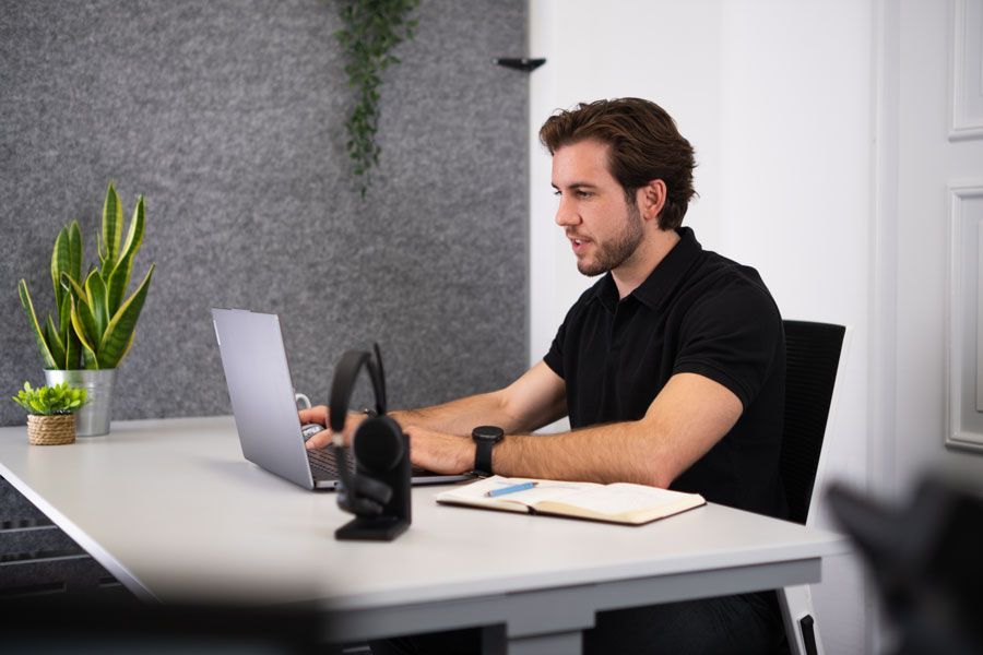Ein Mann arbeitet an einem Laptop an einem Schreibtisch in einem modernen Büro; in der Nähe befinden sich ein Notizbuch und Kopfhörer.