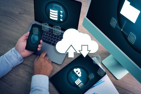 Hände, die Geräte bedienen, umgeben von Symbolen für die Cloud-Synchronisierung auf einem Schreibtisch mit Laptop und Monitoren.