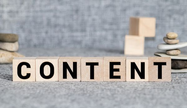 Holzbuchstaben, die das Wort „CONTENT“ bilden, auf grauem Untergrund mit aufgeschichteten Steinen im Hintergrund
Eine Person, die an einem Schreibtisch arbeitet, mit einem Chatbot-KI-Bildschirm und Sprechblasen in einer blauen Büroumgebung.