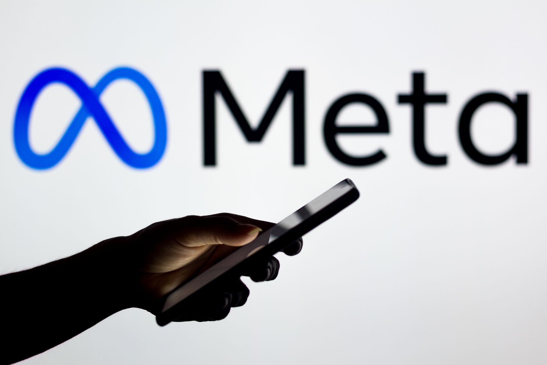 Eine Hand, die ein Smartphone vor dem blauen Meta-Logo auf weißem Hintergrund hält