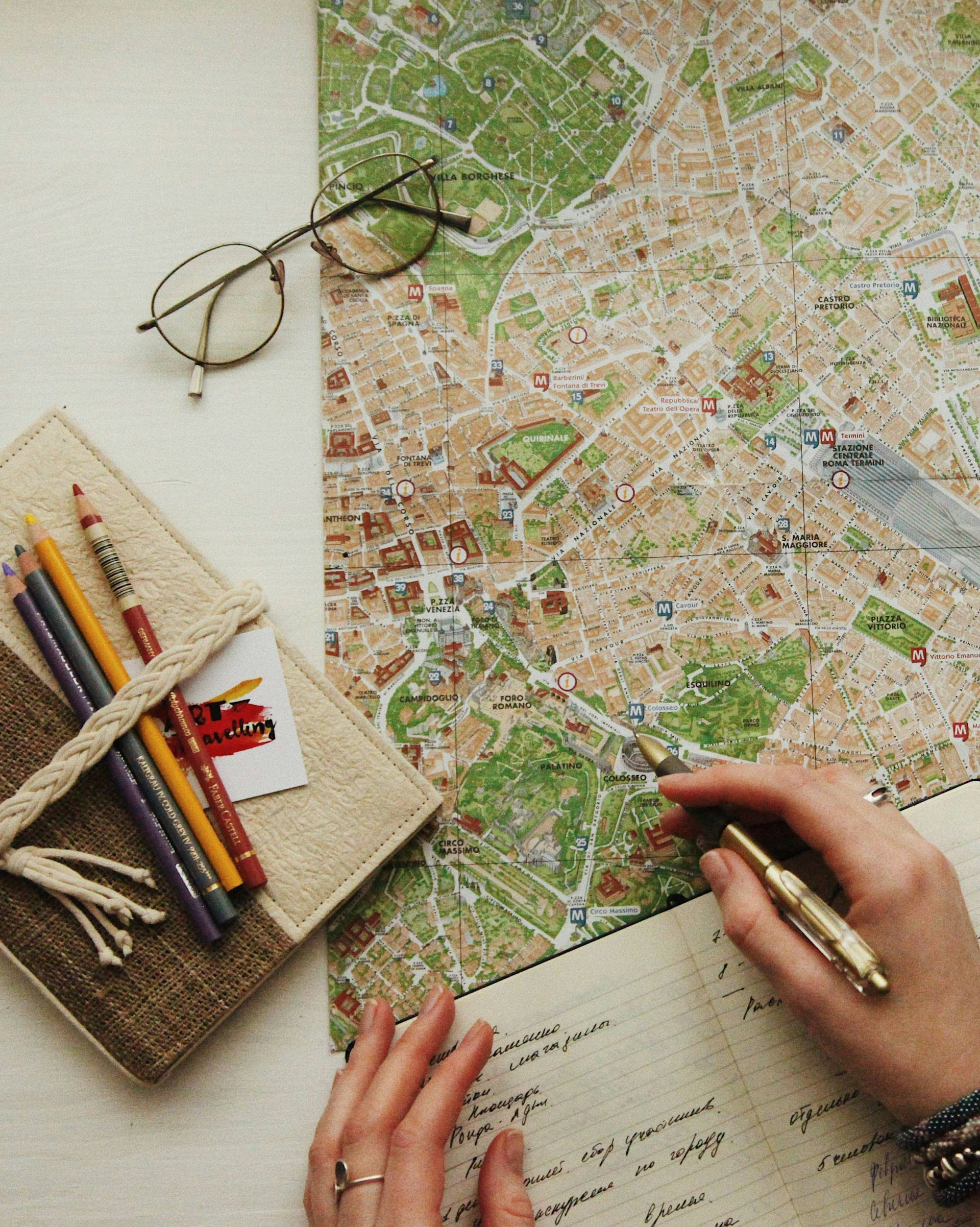 Person plant eine Reise mit Stadtkarte, Notizbuch, Buntstiften und Brille auf einem weissen Schreibtisch.