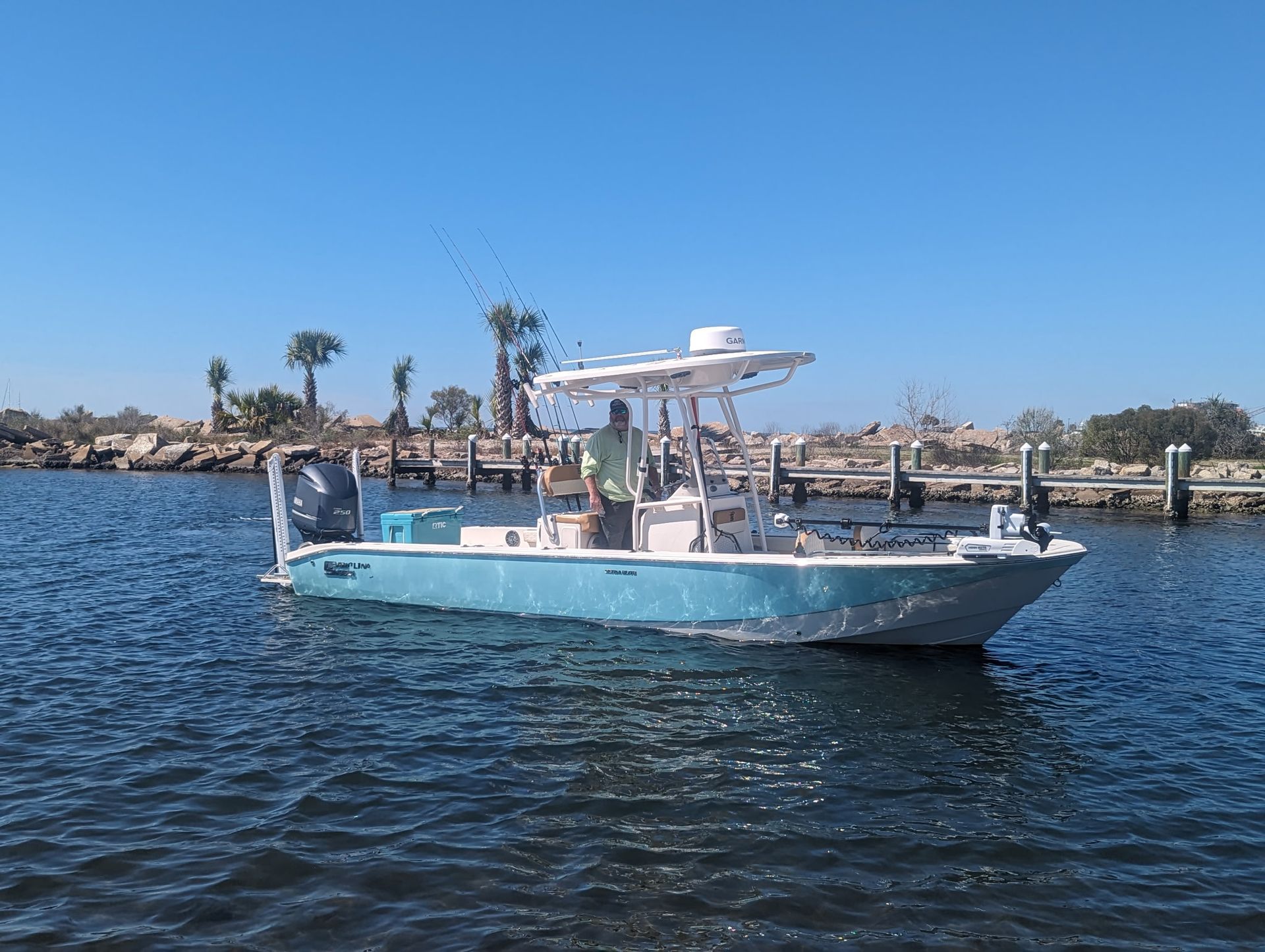 A light blue center console fishing boat cruises through calm water near a shoreline with palm trees and a wooden pier.