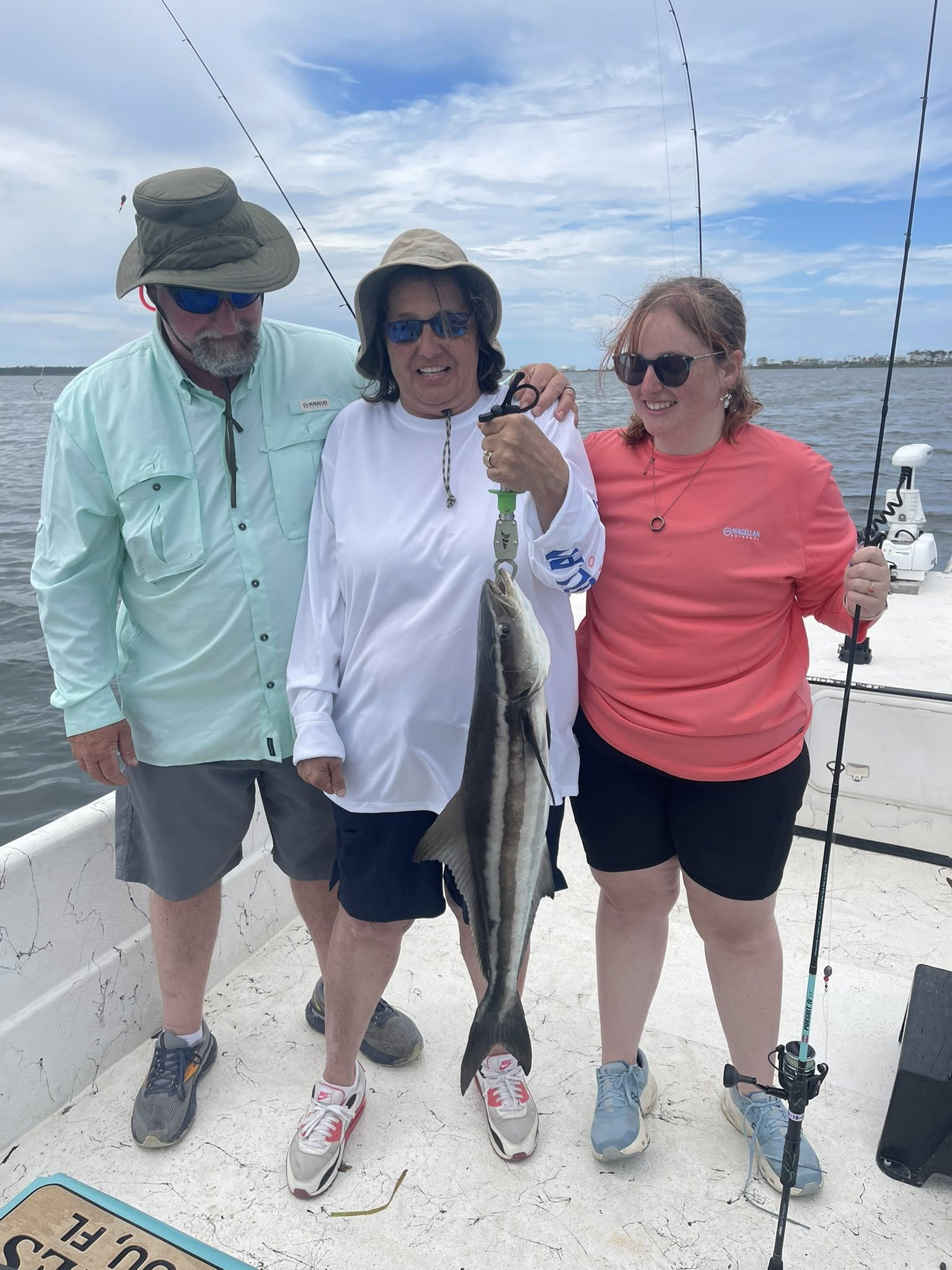 Three people on a boat deck holding a cobia fish, smiling while wearing fishing gear and hats on a sunny day.