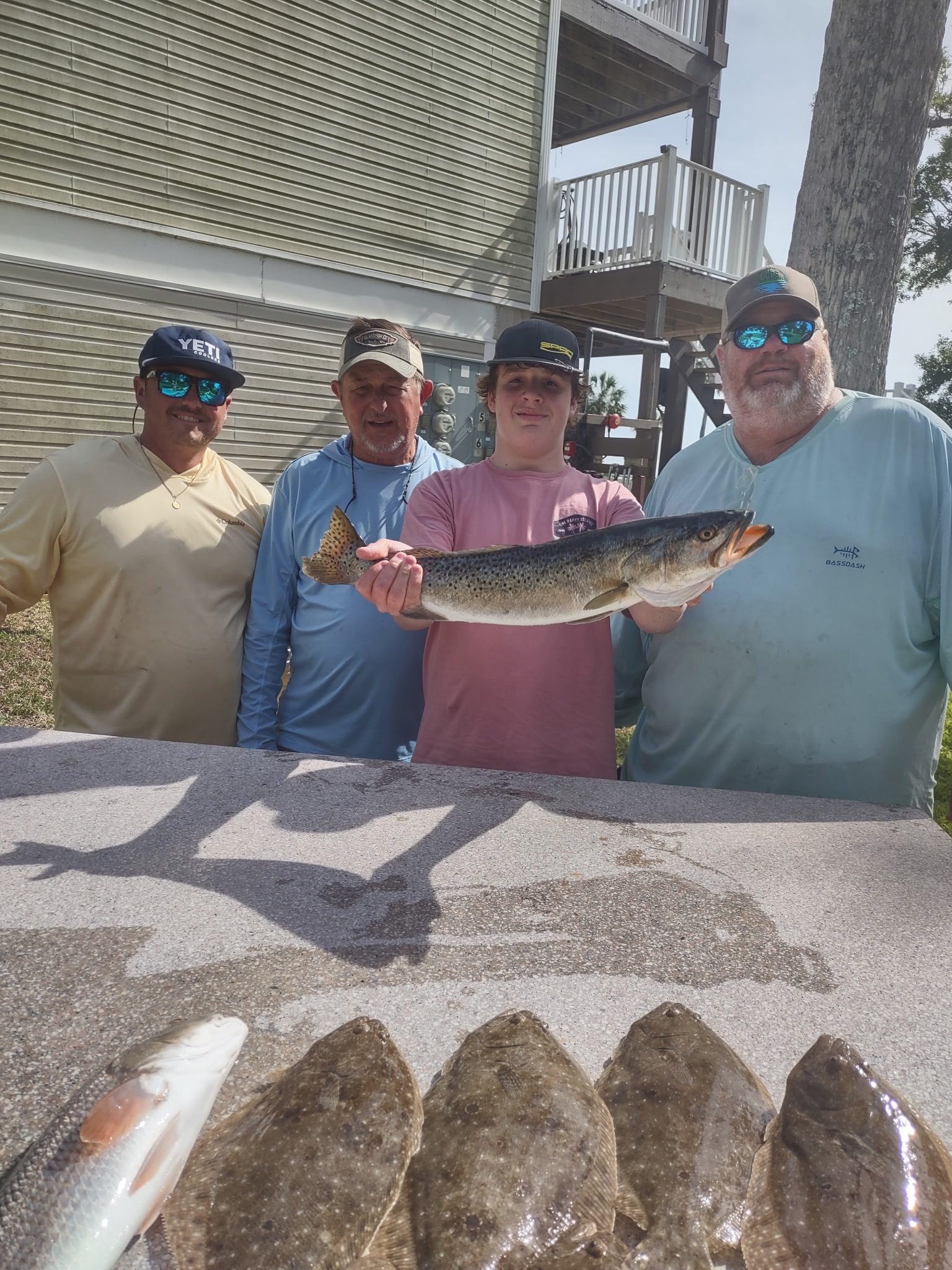 Four people stand in front of a house, holding a large fish over a table with several more fish laid out on it.
