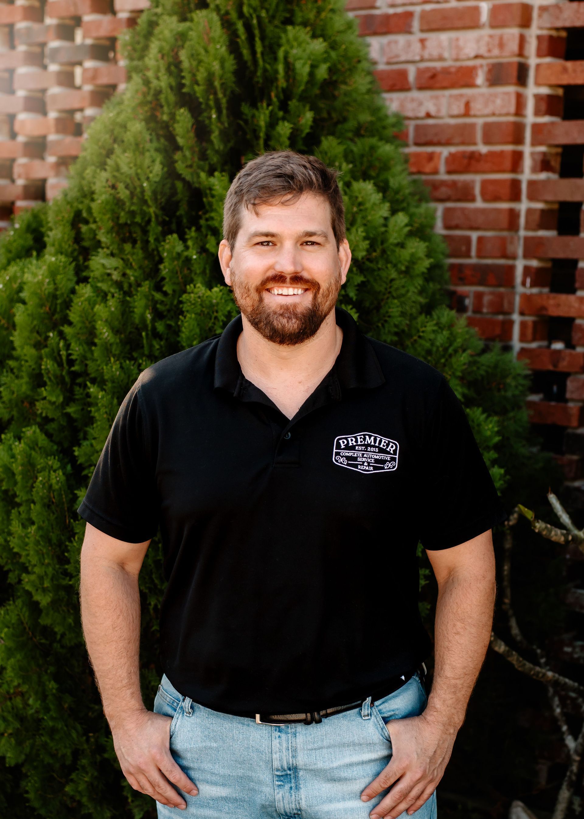 A man in a black shirt and khaki pants is standing in front of a brick wall.